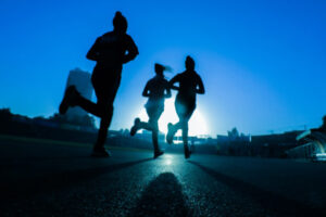 Silhouette of three runners on asphalt running towards sun on horizon against blue sky.