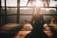 Yoga, which is one of our modalities being done by a woman facing the sun and a palm tree in the distance